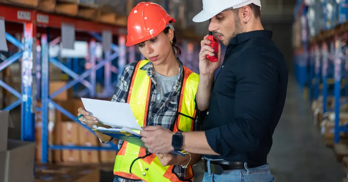 Two warehouse workers in hard hats checking a shipment list between pallet racks, one on a handheld radio